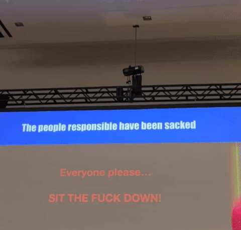 a beach ball flies by a screen that says 'The people responsible have been sacked. Everyone please... SIT THE FUCK DOWN!'